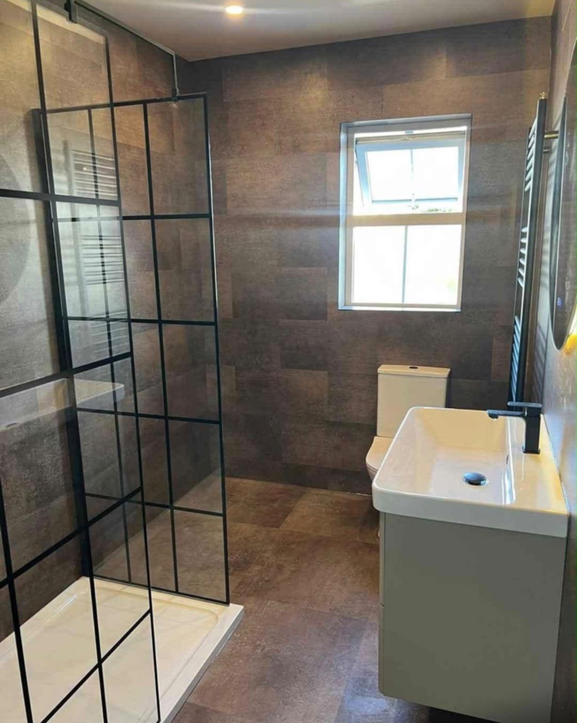 Modern bathroom with walk-in shower, dark tile walls, white vanity sink, window, and heated towel rack
