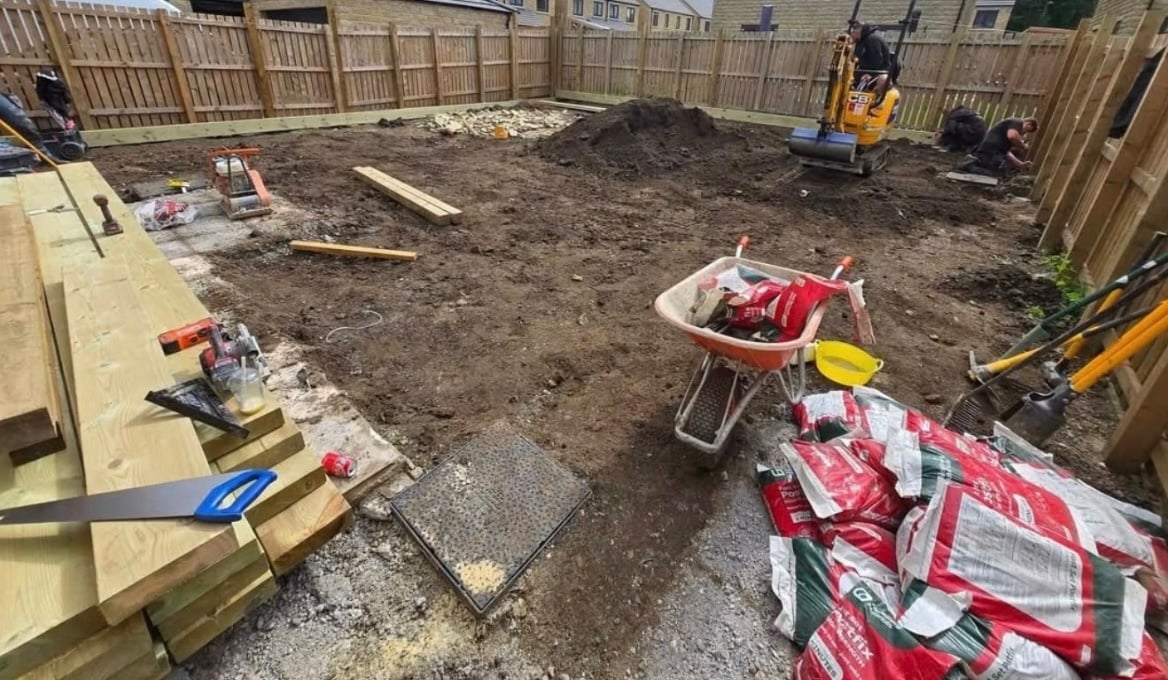Construction site in backyard with wood fencing, dirt pile, wheelbarrow, tools, and building materials stacked on ground
