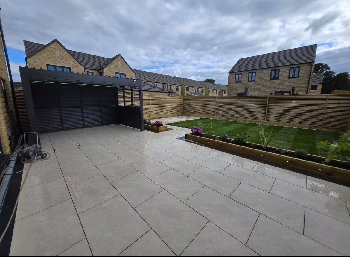 Modern suburban house with large paved patio, black garage, and raised garden beds under cloudy sky