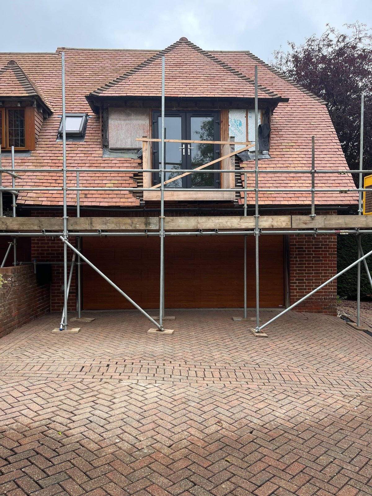 Two-story brick house with terracotta tile roof under renovation, metal scaffolding covering the front facade