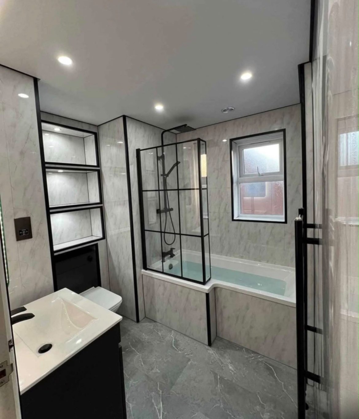 Modern bathroom with marble tiling, black-framed glass shower enclosure, white sink vanity, heated towel rail, and window.